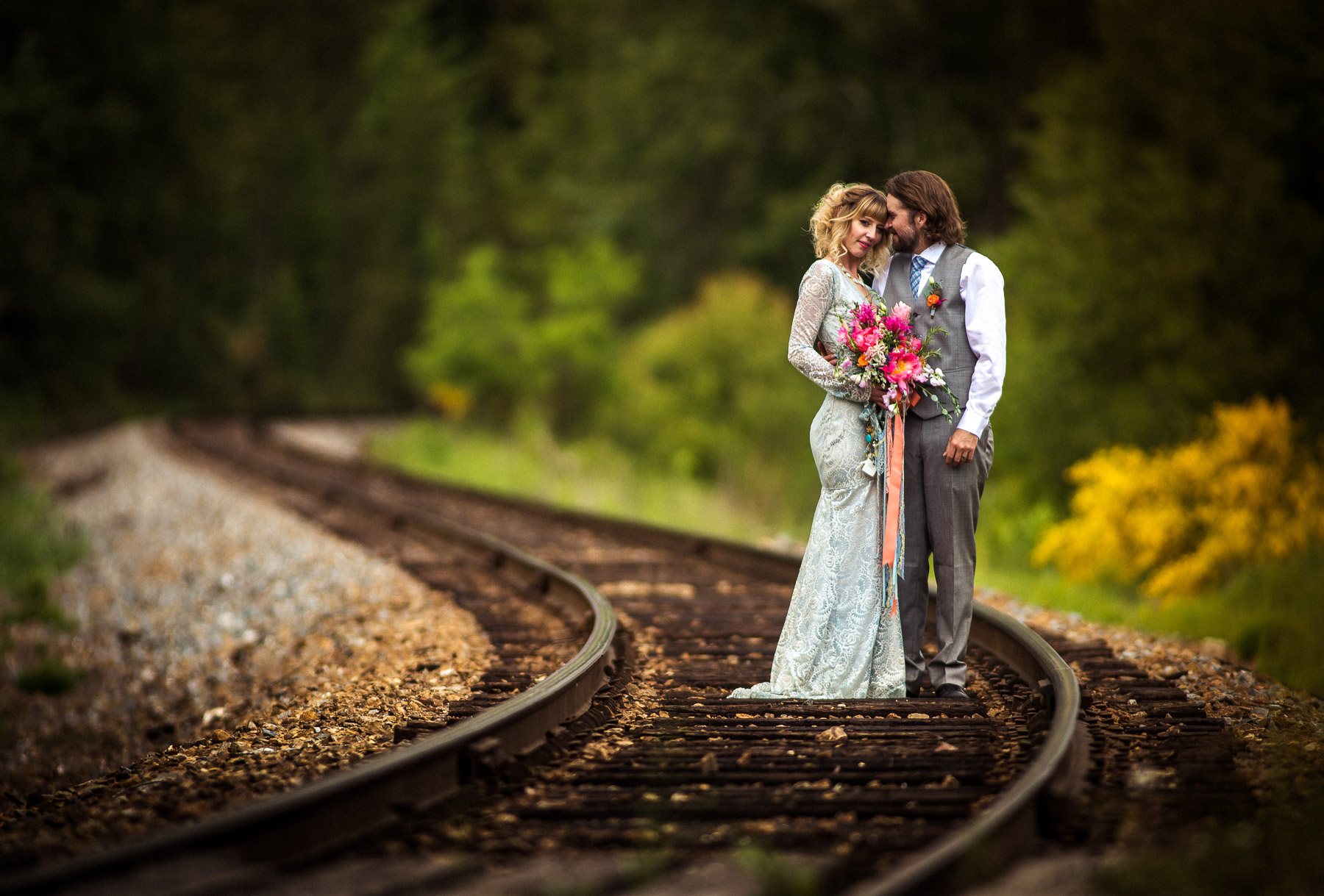 Bride and groom embrace on curved railway tracks, forest lining a soft Kootenay backdrop.